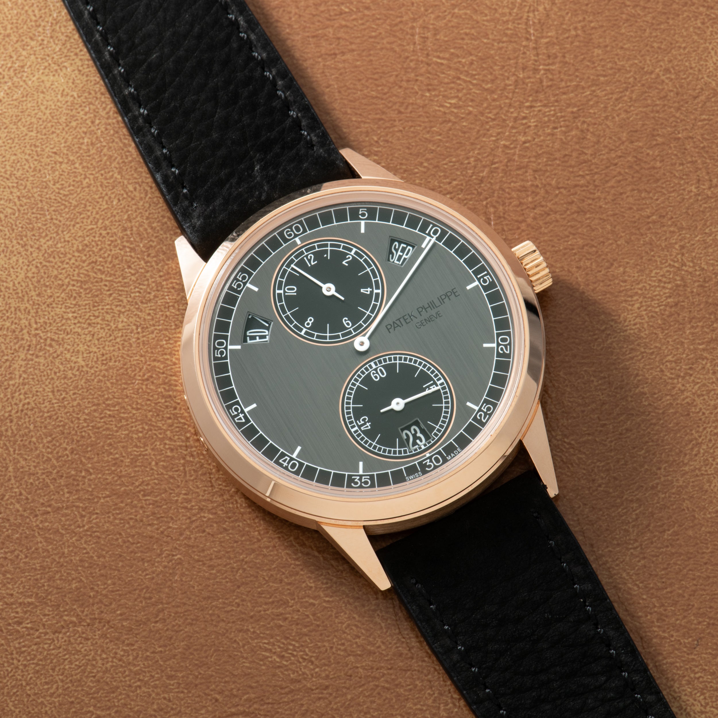 Rose gold watch with gray dial, two subdials, and black leather strap on tan background.