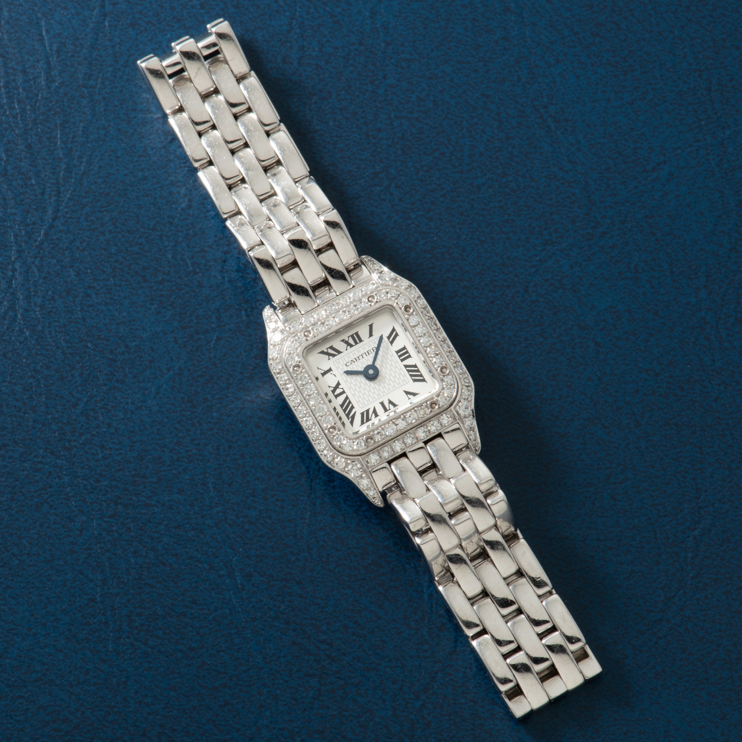 Silver-tone square watch with diamond-set bezel, Roman numeral dial, and multi-link metal bracelet on blue background.