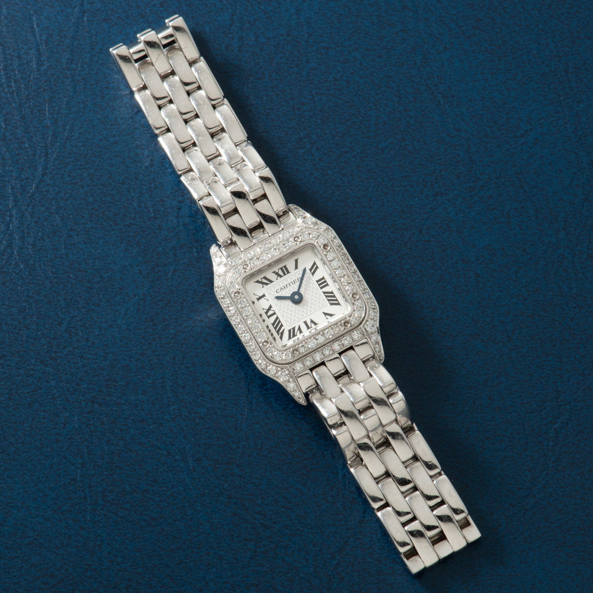 Silver-tone square watch with diamond-set bezel, Roman numeral dial, and multi-link metal bracelet on blue background.