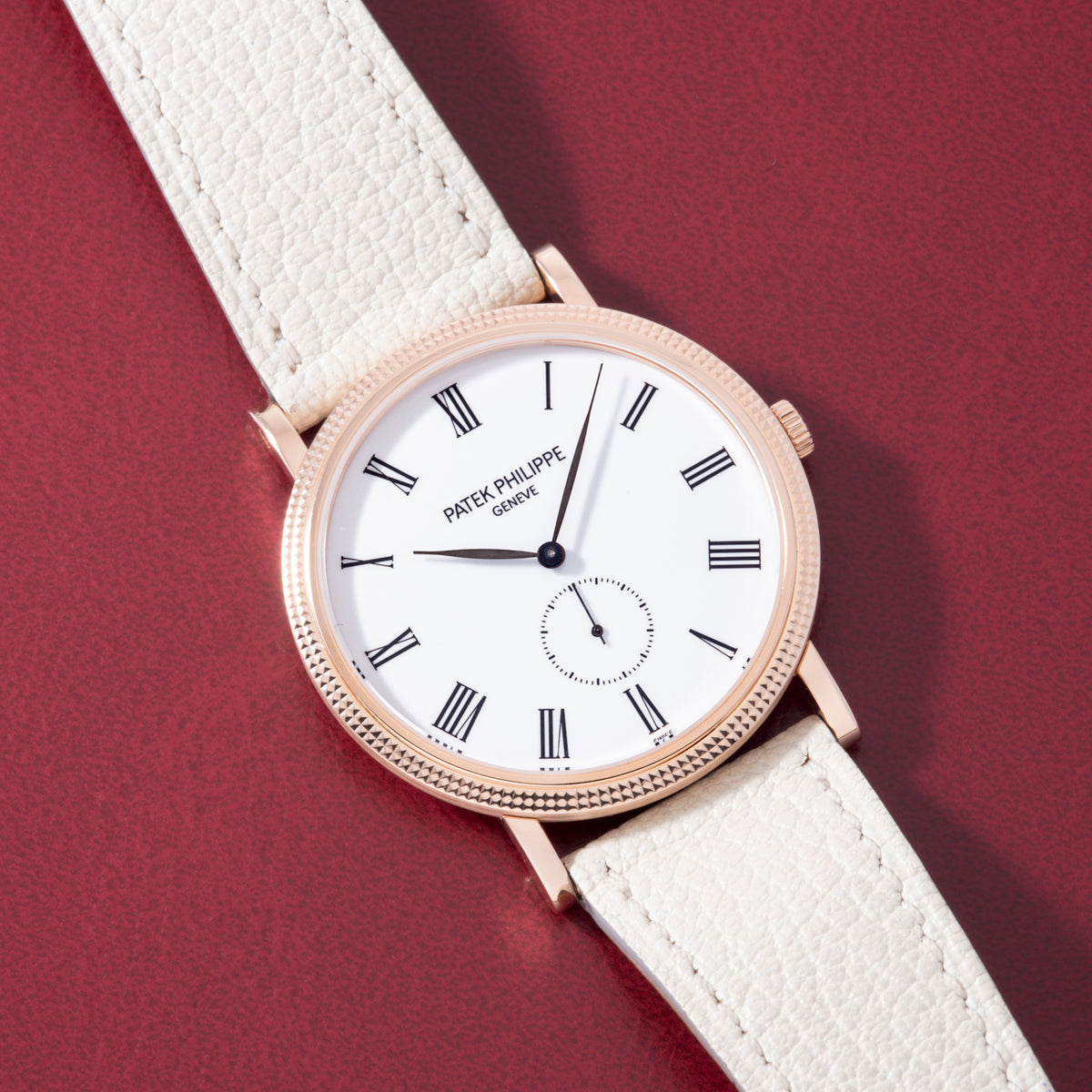 Rose gold watch with white dial, Roman numerals, subdial, and white leather strap on burgundy background.