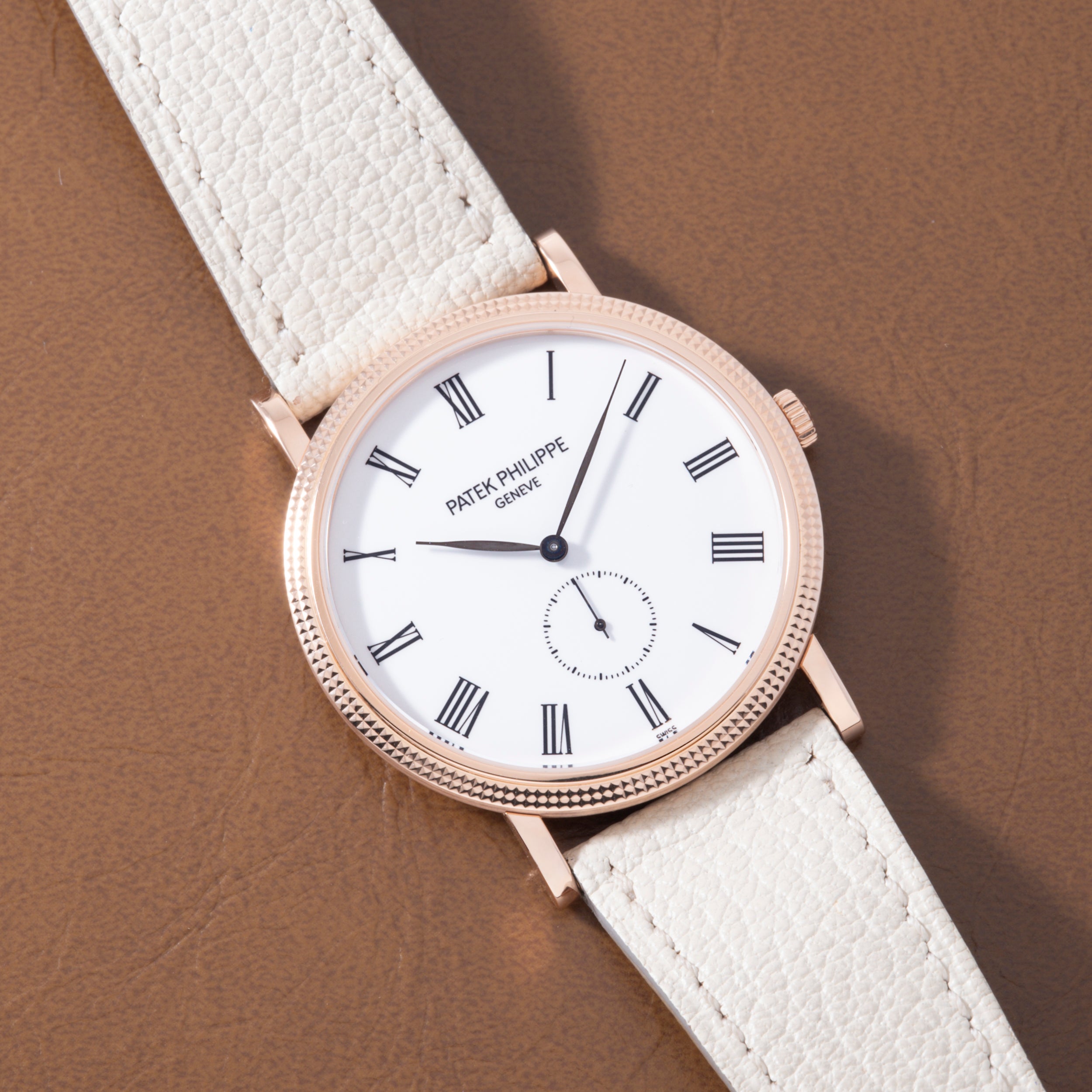 Rose gold watch with white dial, Roman numerals, subdial, and white leather strap on tan background.