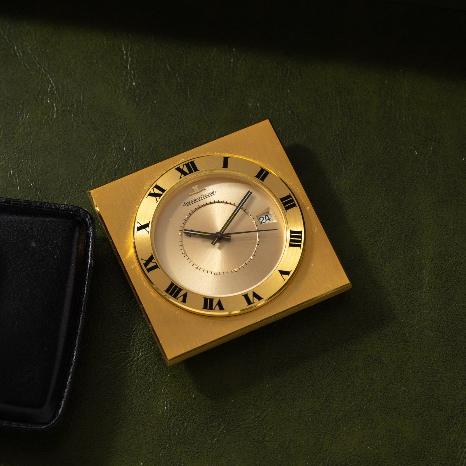 A Jaeger LeCoultre Memovox Travel Calendar Alarm Clock with black Roman numerals and a date window rests on green leather beside a black case, displaying approximately 1:51.