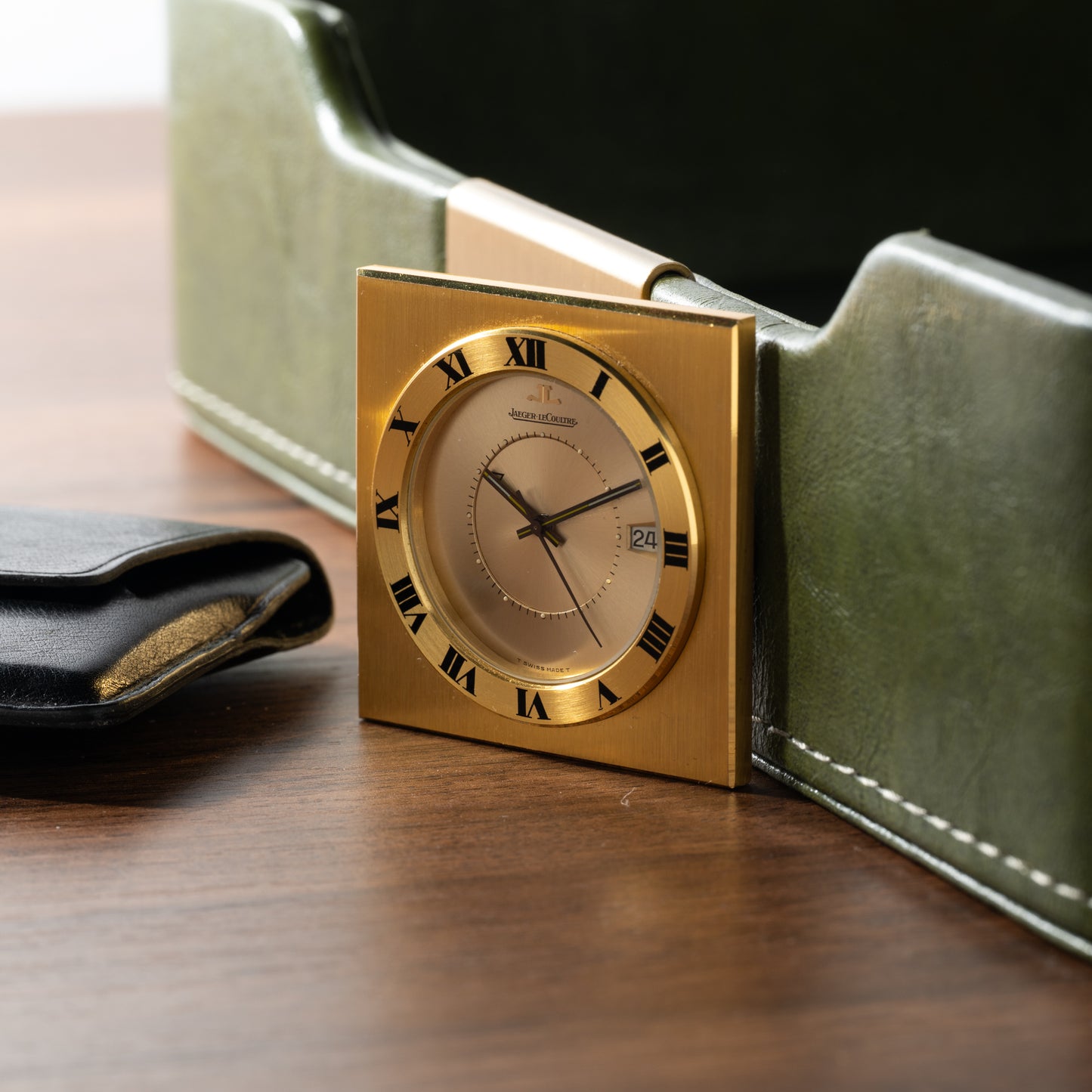 A Jaeger-LeCoultre Memovox Travel Calendar Alarm Clock with Roman numerals sits on a wooden surface beside a black wallet and a green leather holder. The clock, by Jaeger LeCoultre, shows 2:57 and features a date window.
