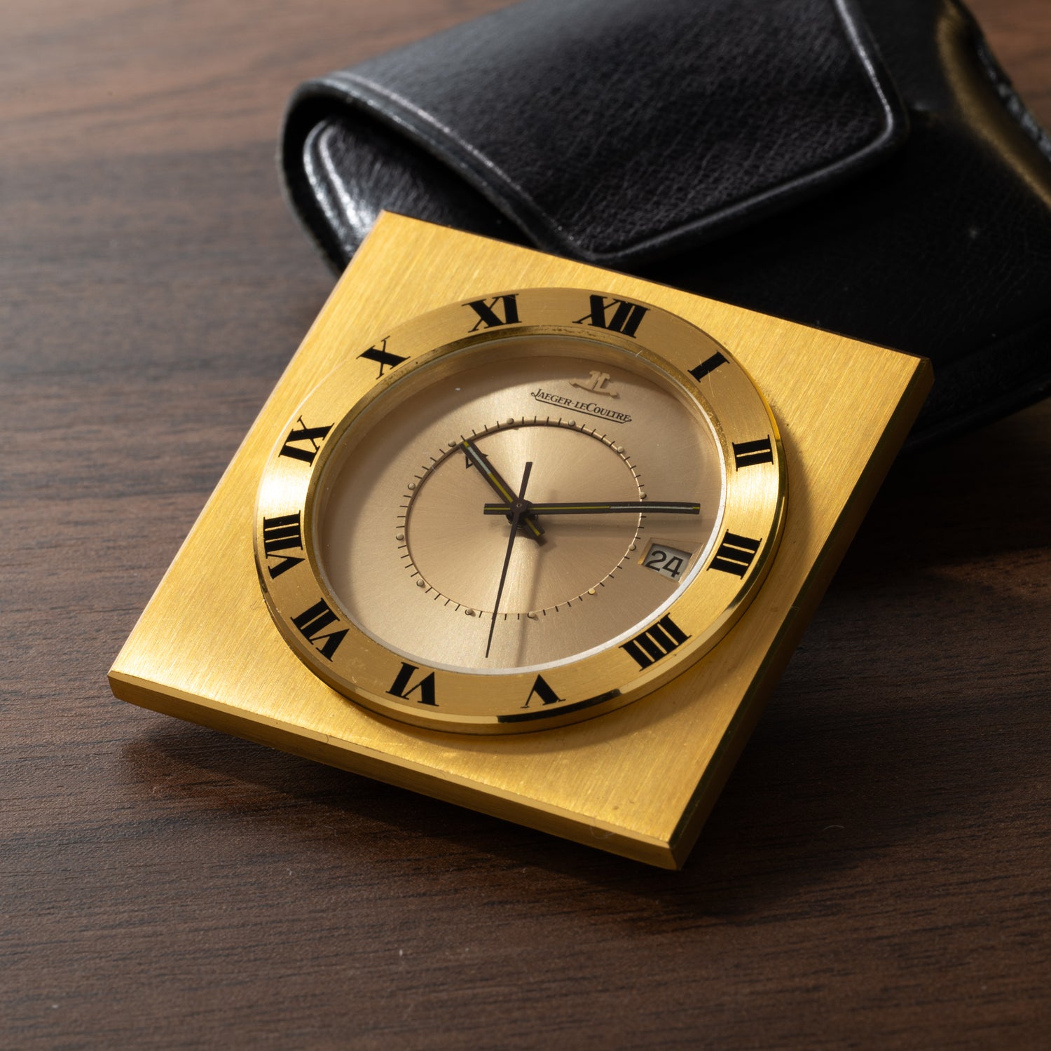 A square, gilt-brass Jaeger LeCoultre Memovox Travel Calendar Alarm Clock with Roman numerals and black hands sits on a wooden surface next to its black leather case, displaying the time and a date window.