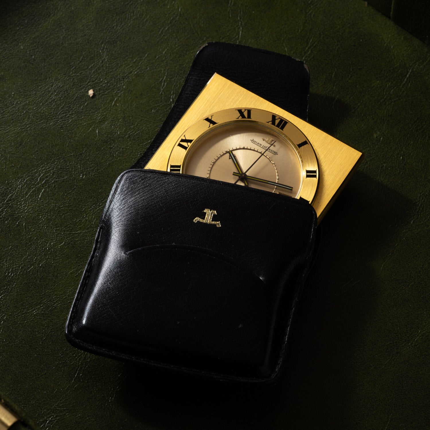 A Jaeger LeCoultre Memovox Travel Calendar Alarm Clock with Roman numerals, partly nestled in a black leather logo-embossed case, sits on a dark green surface.