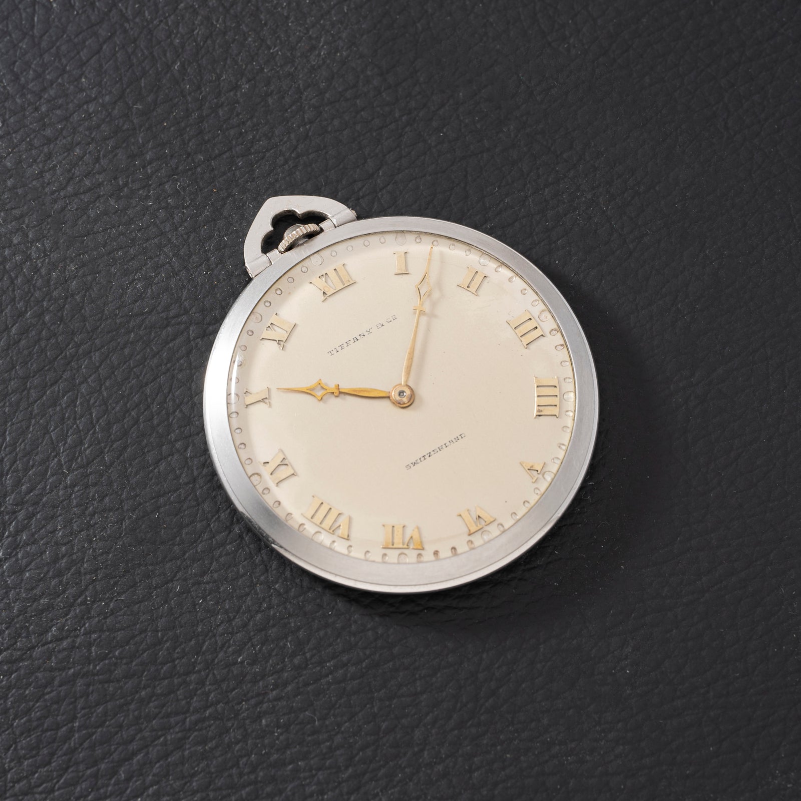 The Tiffany & Co. Platinum Pocket Watch, featuring gold Roman numerals and hands, rests on a textured black surface at 6:17, showcasing the brand's classic mechanical craftsmanship.