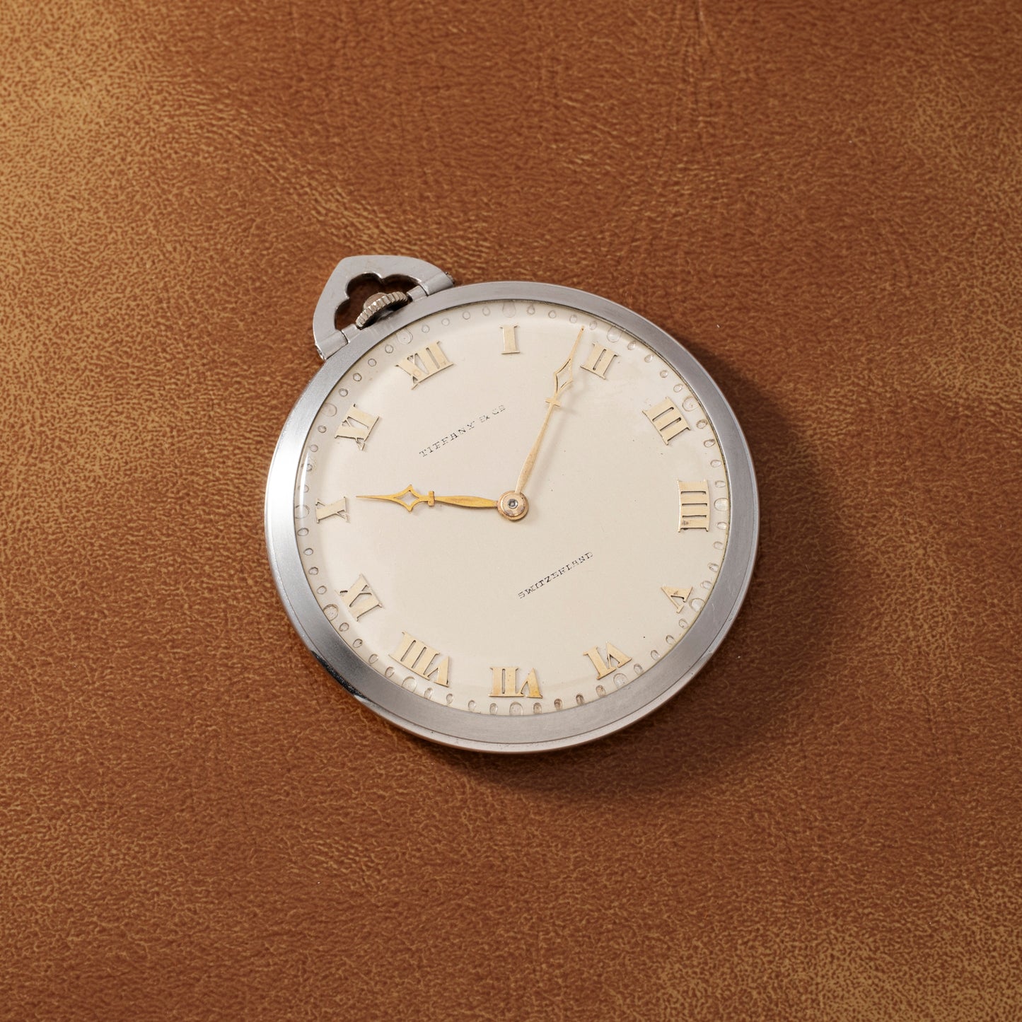 A Tiffany & Co. Platinum Pocket Watch with gold hands and Roman numerals rests on a brown leather surface, the watch face displaying the brand name.