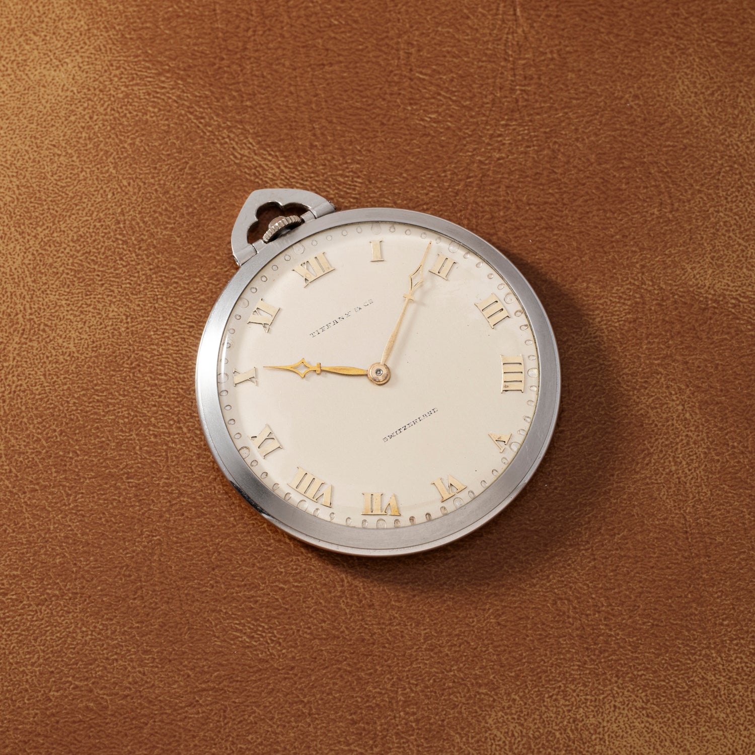 A Tiffany & Co. Platinum Pocket Watch with gold hands and Roman numerals rests on a brown leather surface, the watch face displaying the brand name.