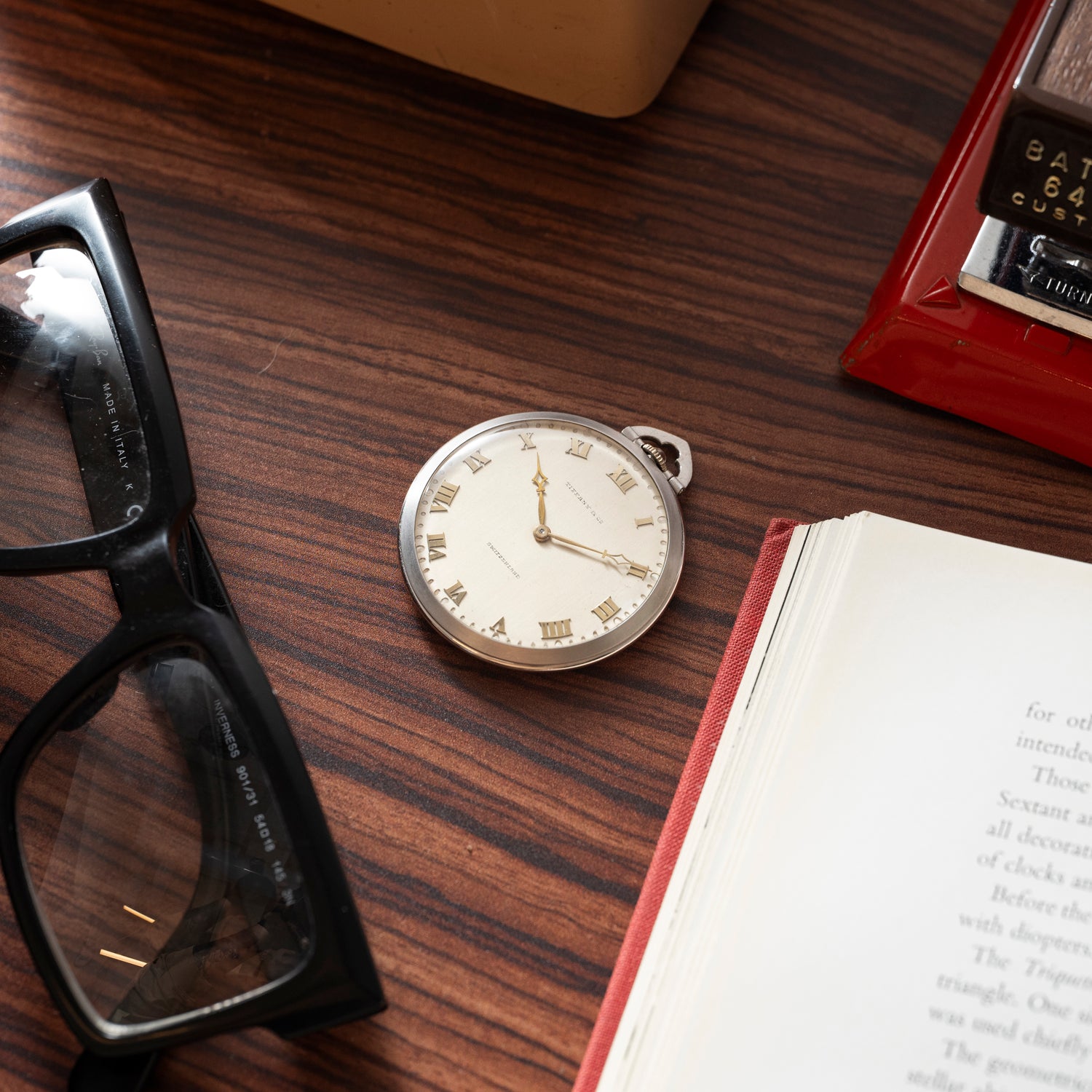 A Tiffany & Co. Platinum Pocket Watch with Roman numerals rests on a wooden table beside black glasses, an open book, and a red box.