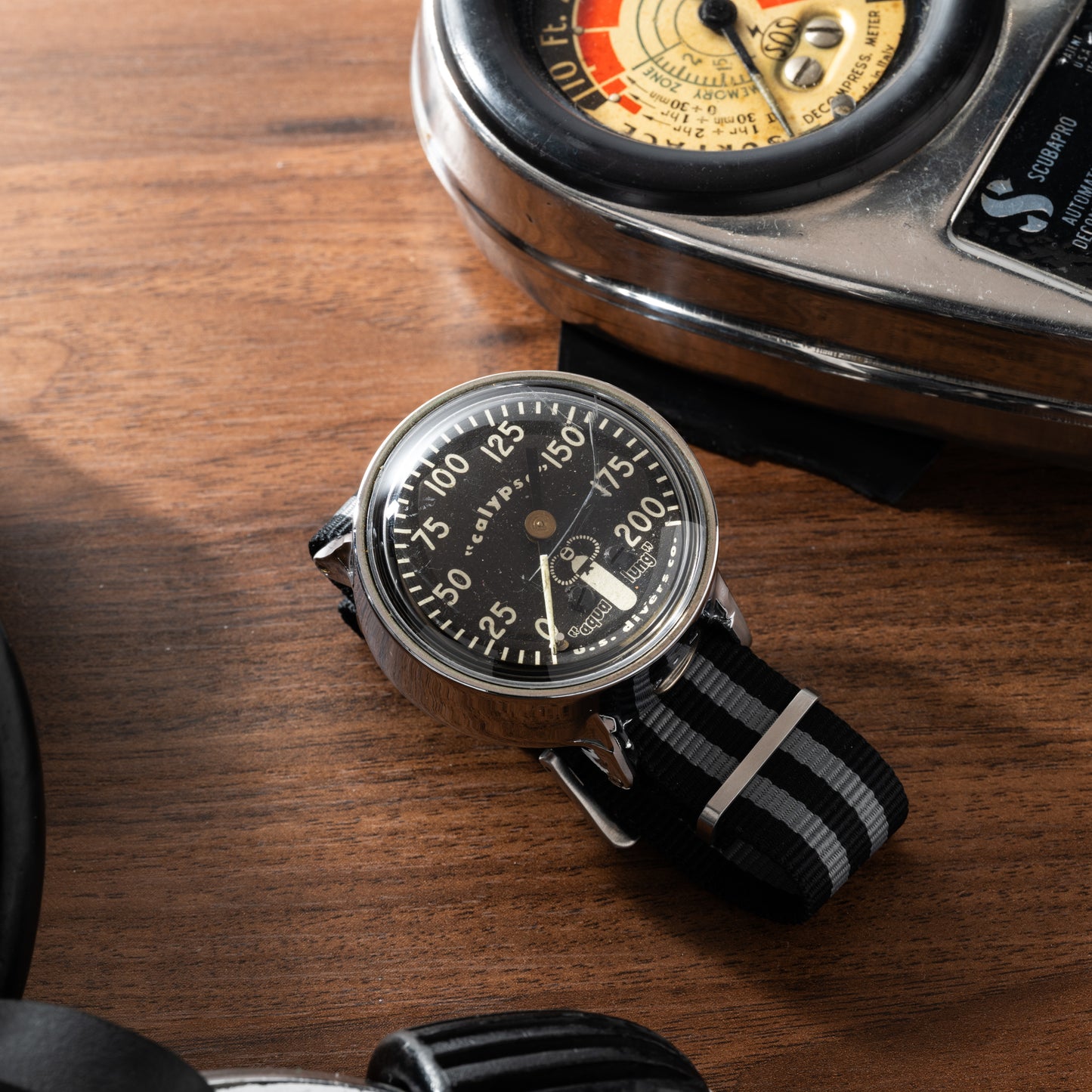 A black and silver Calypso Depth Gauge by Calypso, featuring a striped strap, sits on wood beside a vintage device with dials, evoking classic scuba diving adventures.
