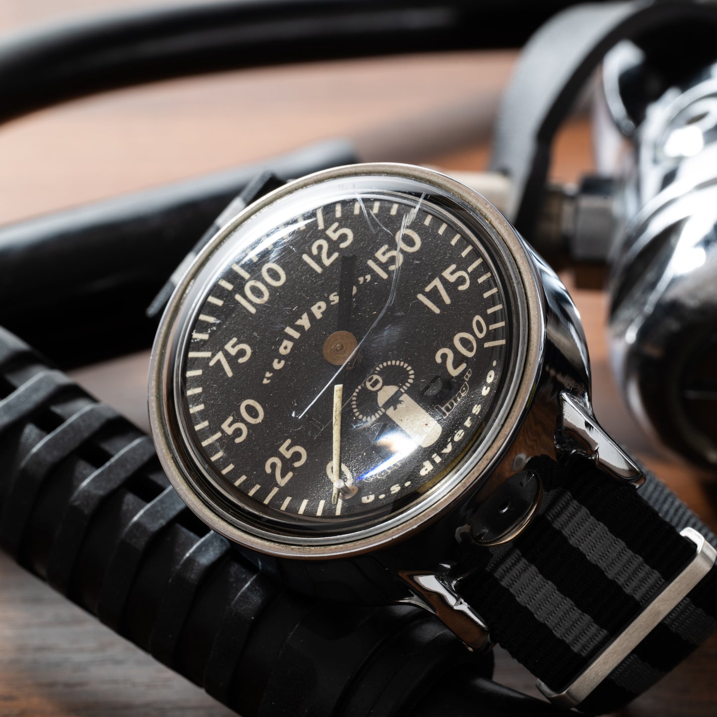 A vintage dive watch with a black dial and striped nylon strap rests on diving gear next to a Calypso Depth Gauge by Calypso, both placed on a wooden surface.