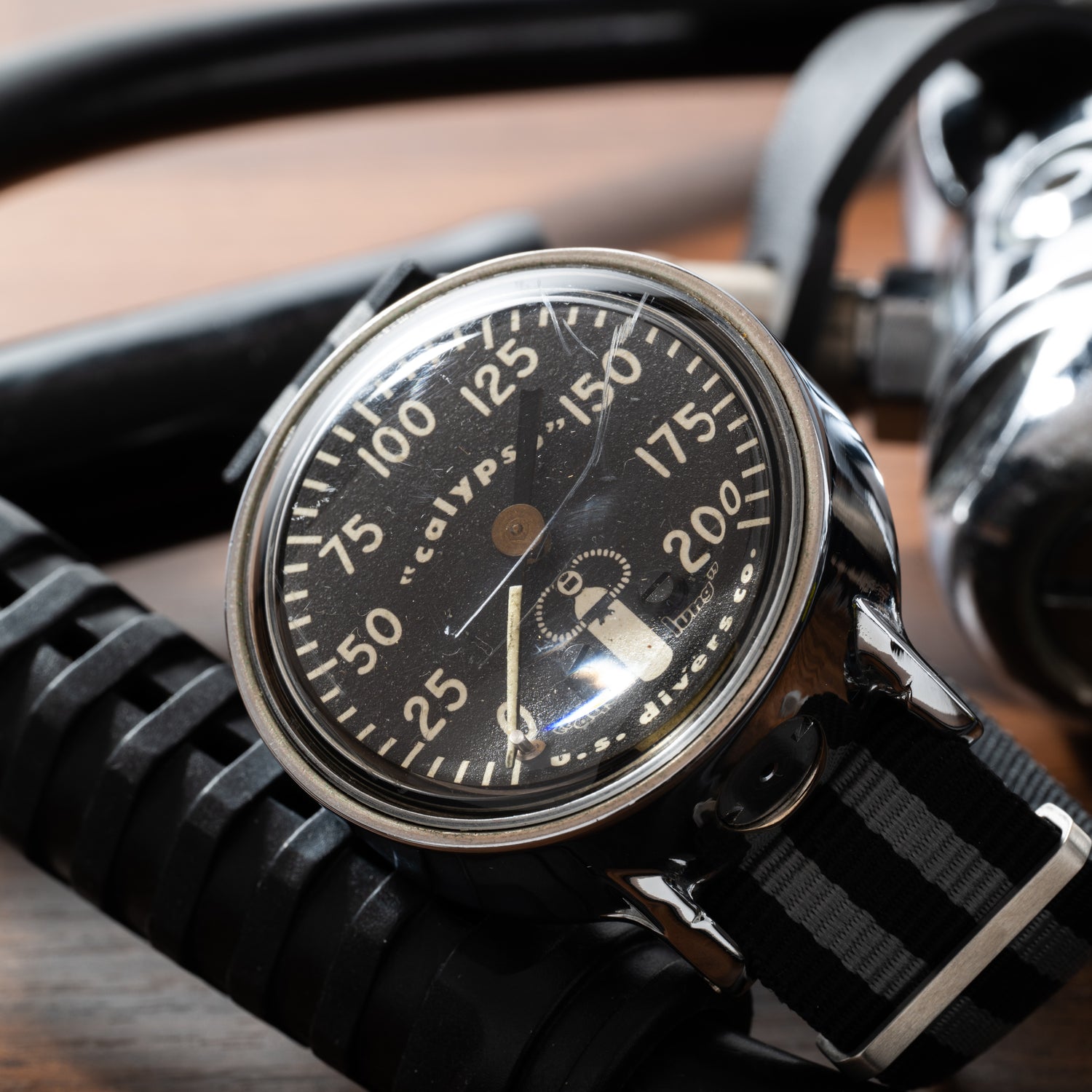 A vintage dive watch with a black dial and striped nylon strap rests on diving gear next to a Calypso Depth Gauge by Calypso, both placed on a wooden surface.