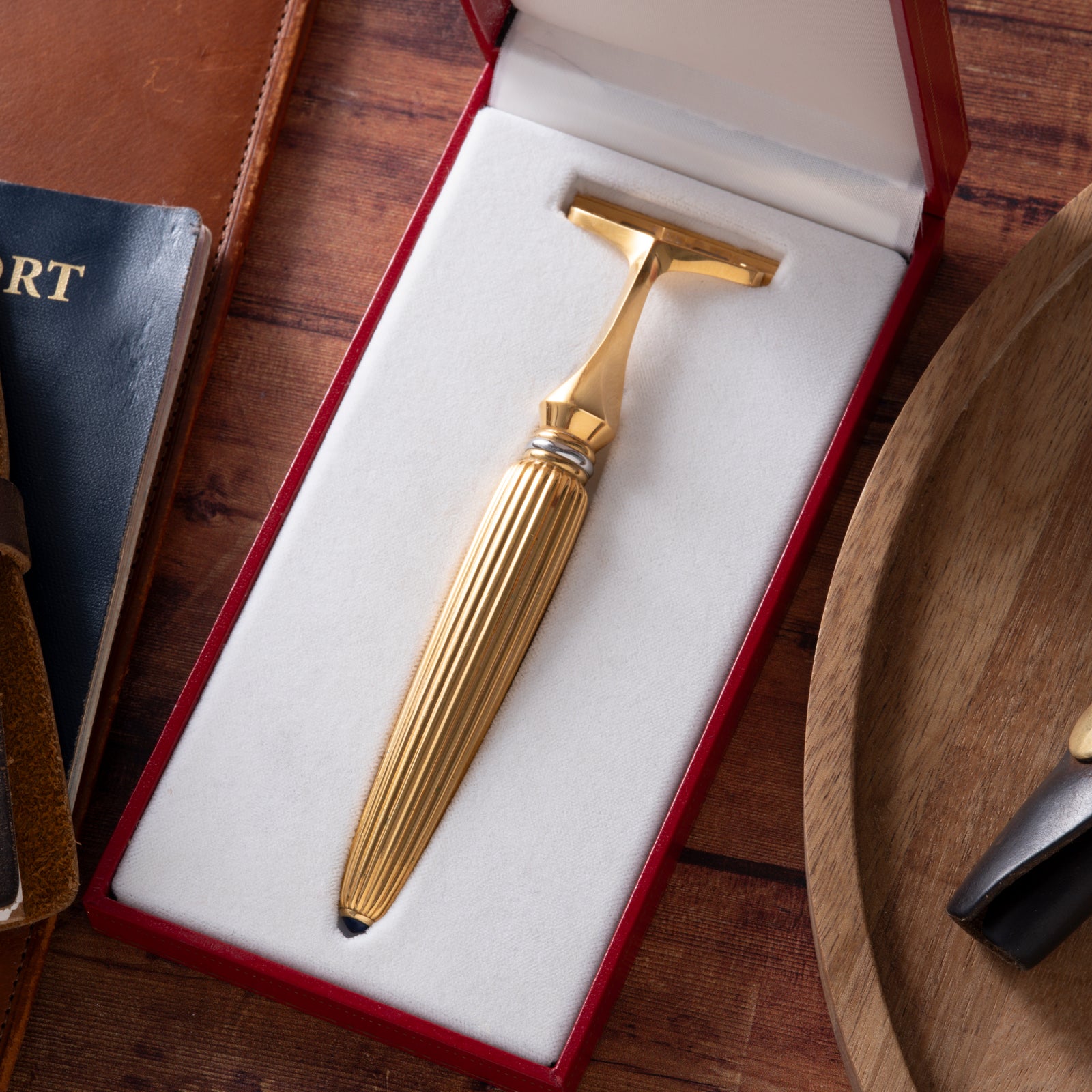 The Cartier Pasha de Cartier Razor by Cartier features an 18K gold-plated, textured handle and is presented in a red and white box on a wooden surface beside a notebook and round wooden tray.