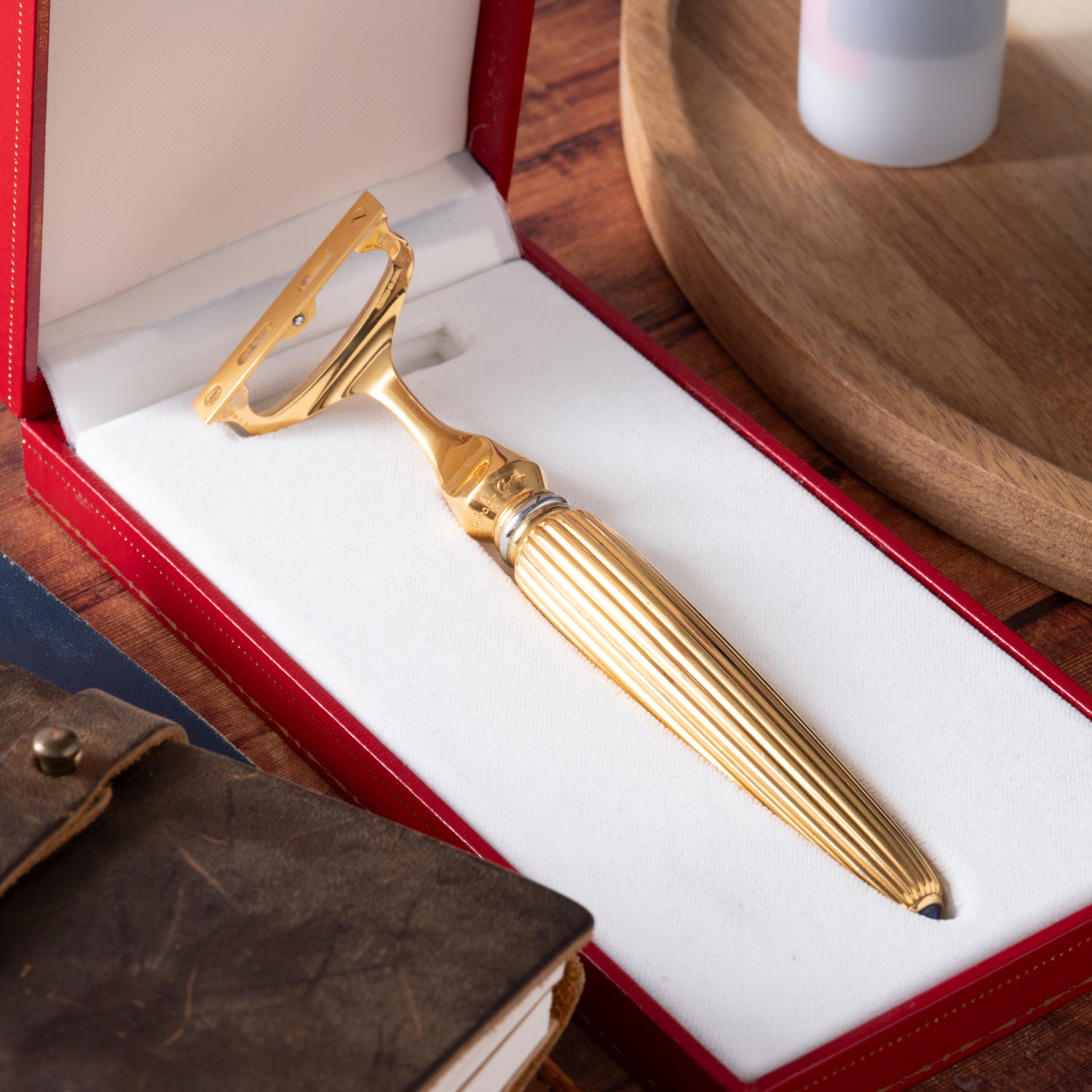 The Cartier Pasha de Cartier Razor by Cartier, featuring a textured handle, sits in a white and red display case on a wooden surface beside a leather-bound notebook and a round wooden tray.