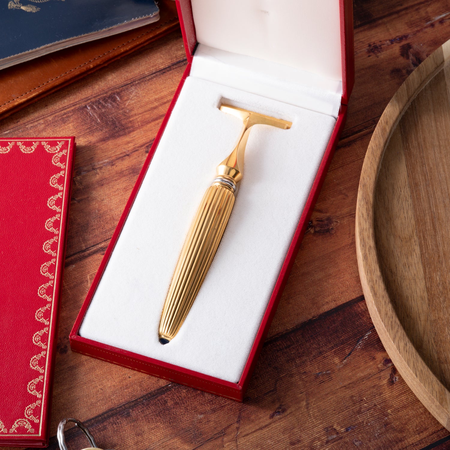 The Cartier Pasha de Cartier Razor by Cartier, featuring an 18K gold-plated ribbed handle, is displayed in a white-cushioned red box on a wooden table near a brown tray, red book, and other objects.