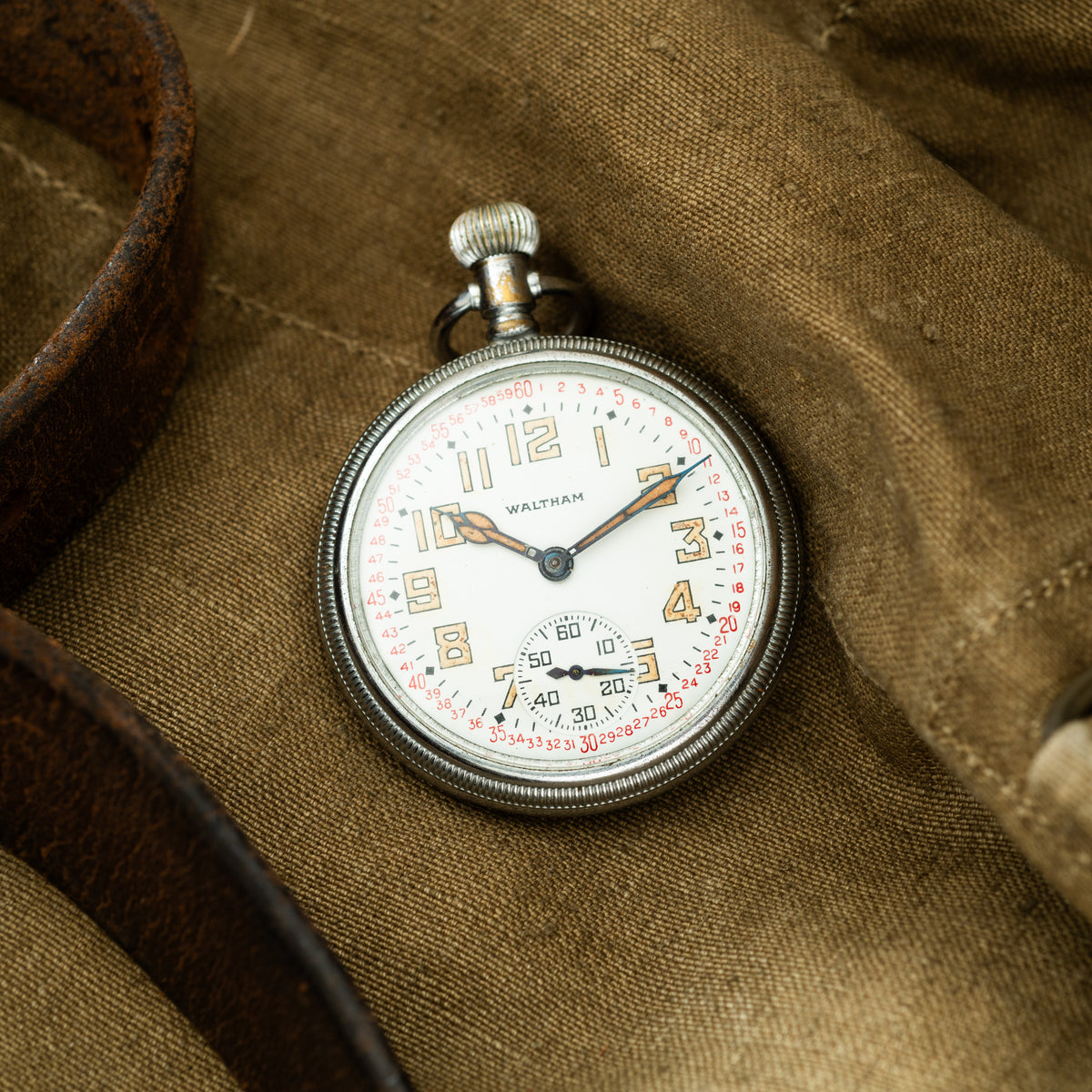 Waltham pocket watch with white dial, Arabic numerals, subsidiary seconds dial, and metal case on khaki fabric.