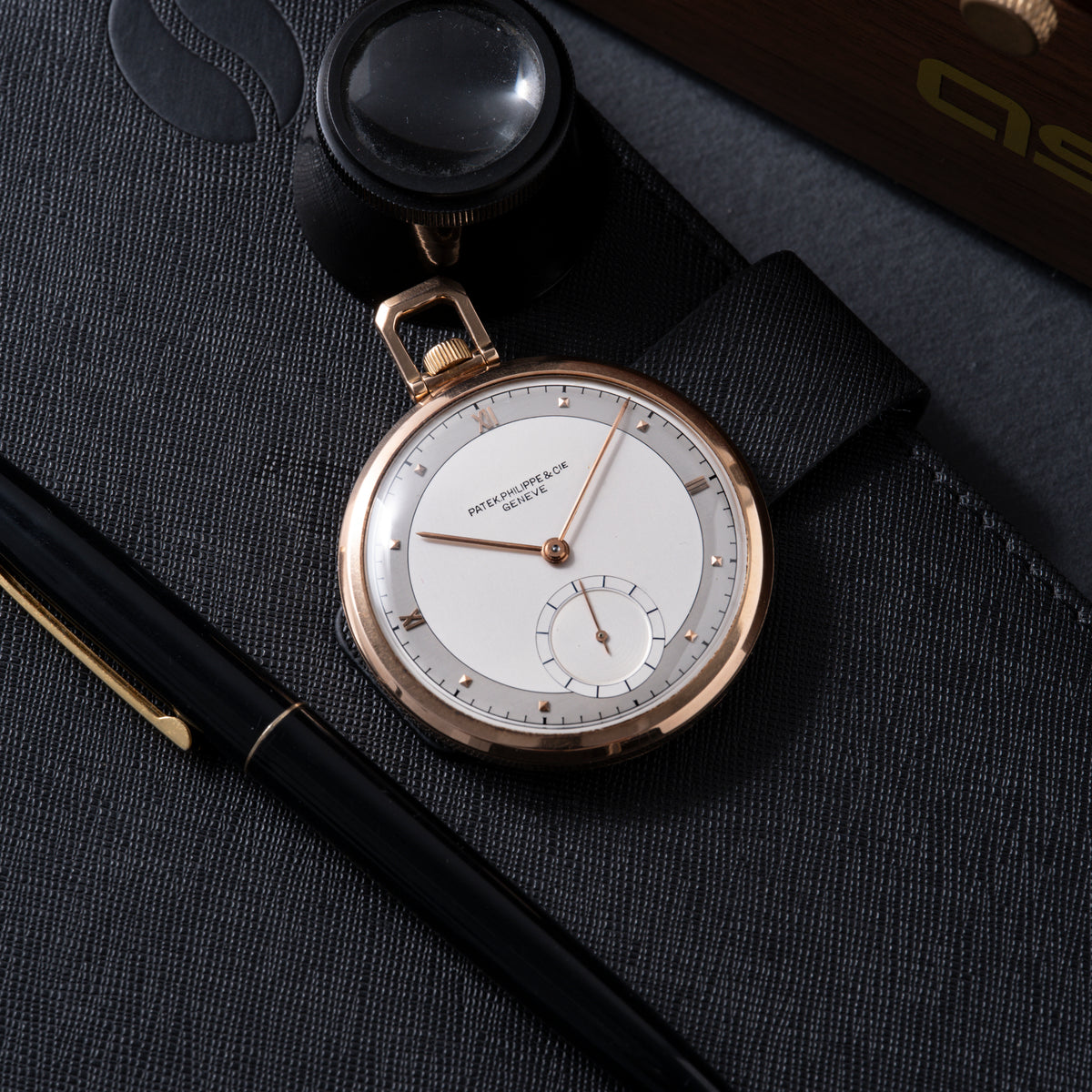 Rose gold pocket watch with white dial, small seconds subdial, and black leather chain on gray fabric surface.