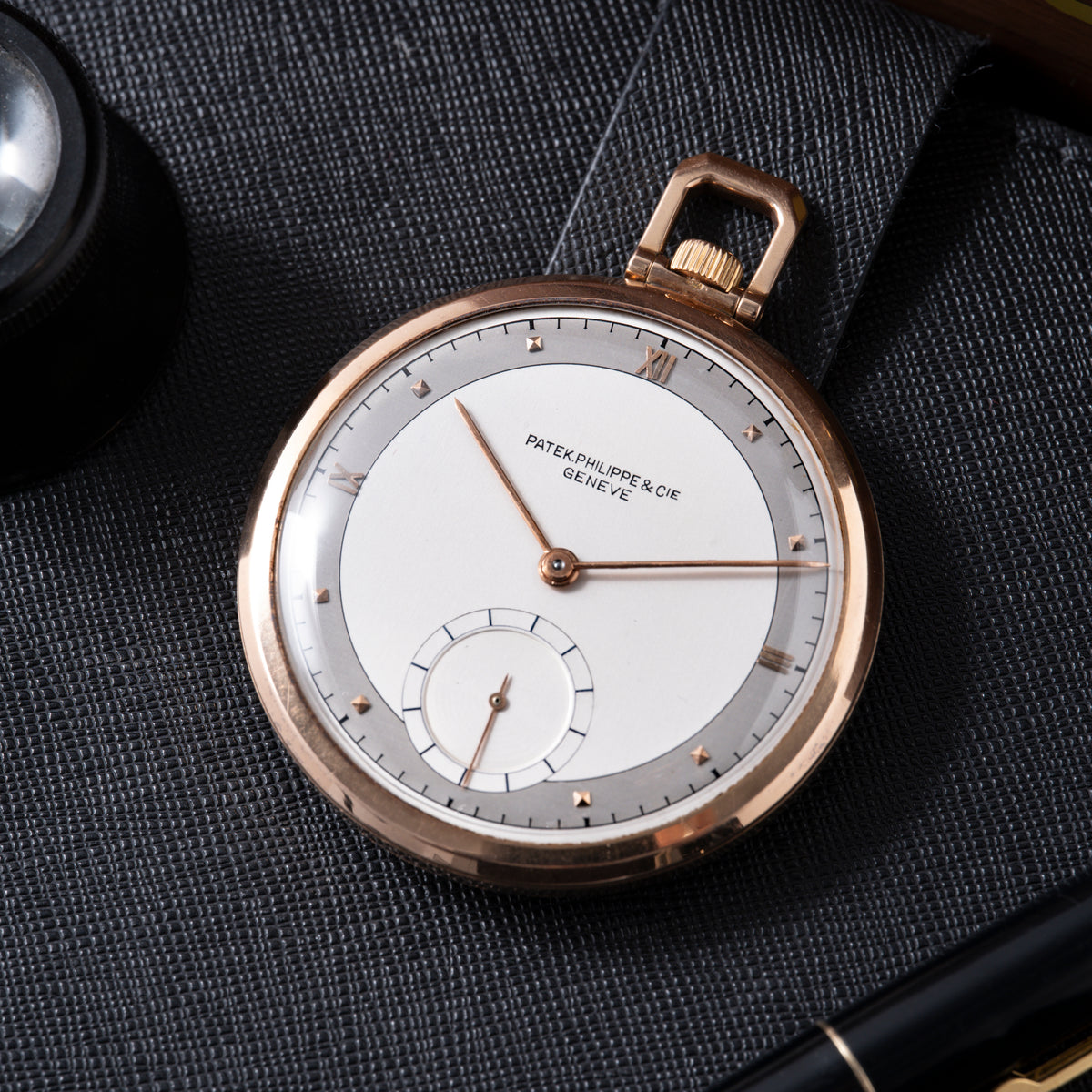 Rose gold pocket watch with white dial, Roman numerals, and subsidiary seconds dial on dark textured fabric.