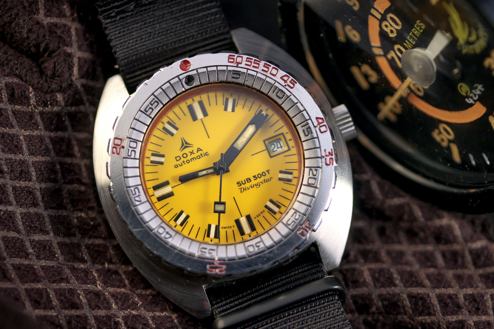 A close-up of the DOXA Sub 300T Divingstar by DOXA, featuring a bright yellow dial, black strap, and silver bezel. The watch shows the date and is placed on textured brown fabric beside another dial.