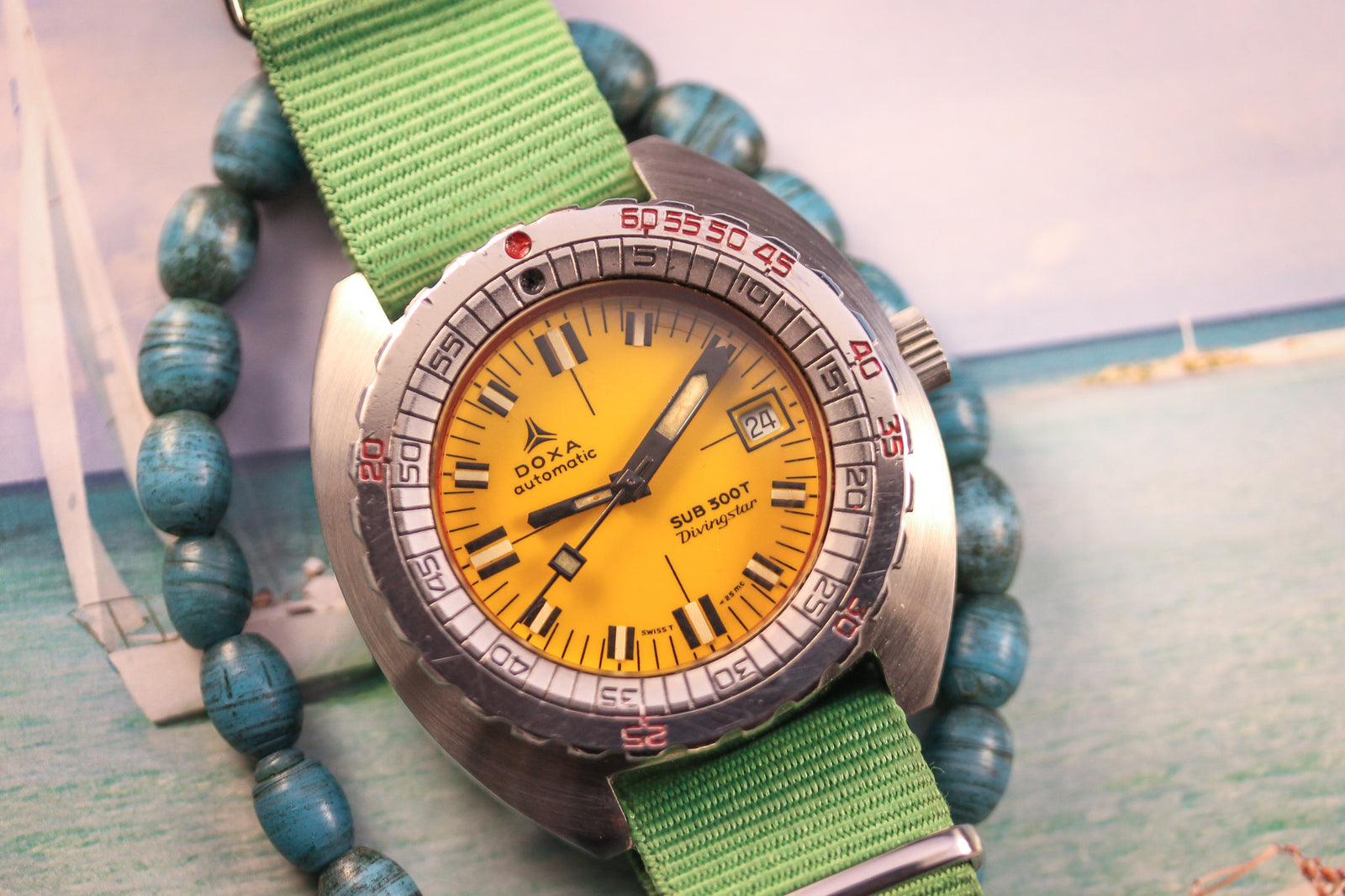 A close-up of the DOXA Sub 300T Divingstar from DOXA, featuring a yellow dial, silver bezel, and green strap, resting near turquoise beads with part of a sailboat photo in the background.