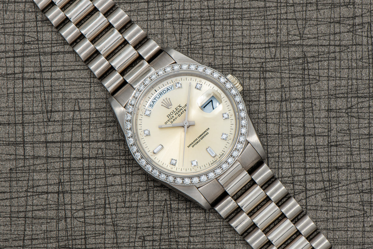 Silver-tone watch with cream dial, diamond bezel, day-date window, and matching metal bracelet on gray textured background.