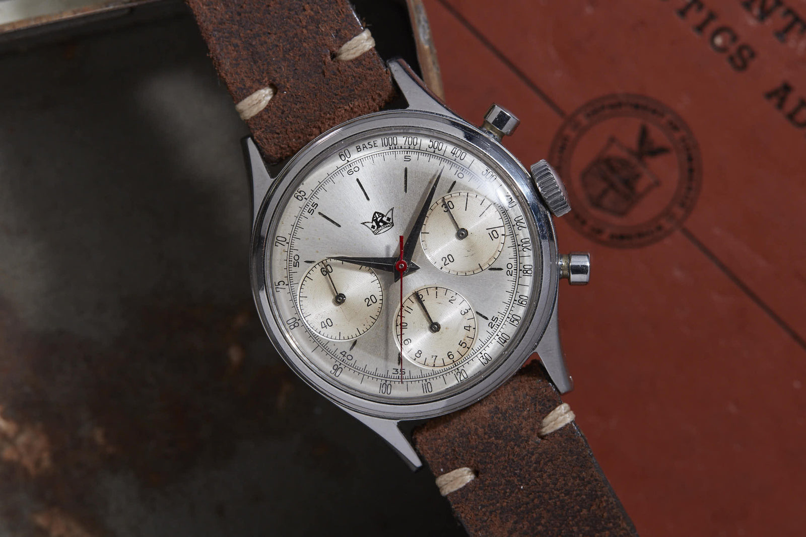A Breitling Chronograph wristwatch with a silver face, three subdials, brown leather strap, and red seconds hand rests near a red booklet—a classic collector's piece from Breitling.