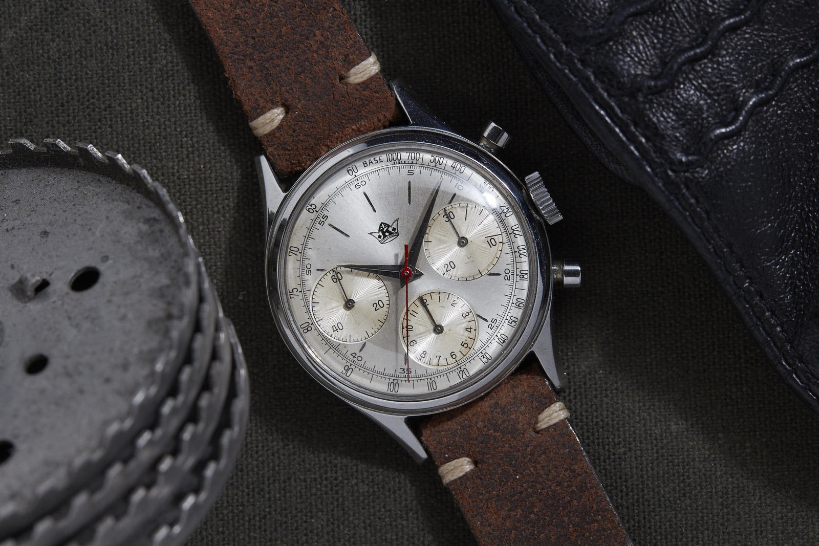 A Breitling Chronograph wristwatch with a brown leather strap and silver dial rests on dark fabric beside a black leather case and a perforated metal object, embodying the timeless appeal of classic Breitling watches.