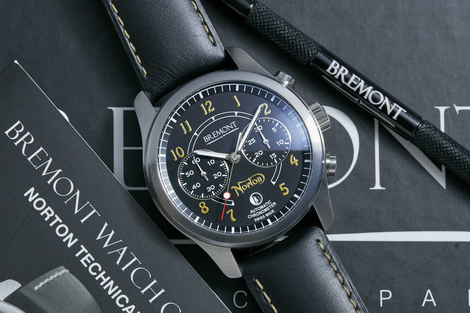 The Bremont Norton Limited Edition Chronograph, featuring a black dial with yellow numerals and a leather strap, is shown with a Bremont technical manual and a branded tool.