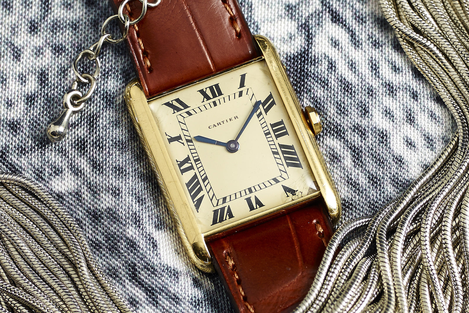 A Cartier Tank Louis Manual Wind - 1940s wristwatch in 18k gold with brown leather strap, Roman numerals, and blue hands lies on a patterned fabric decorated with silver chain fringe.