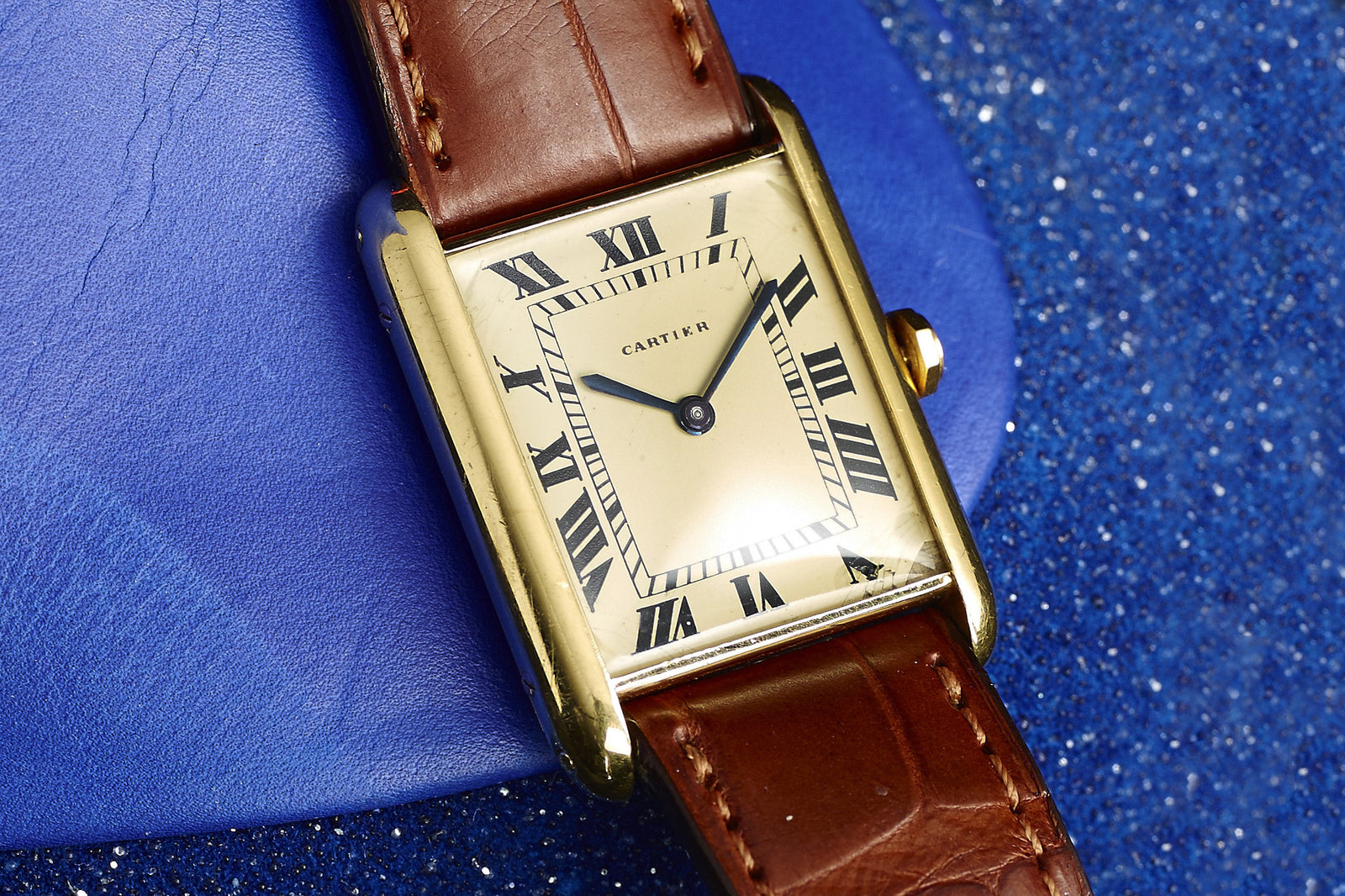 A Cartier Tank Louis Manual Wind -1940s wristwatch with an 18k gold square case, white dial, black Roman numerals, and brown leather strap is shown on a blue textured surface, displaying the time as 10:10.