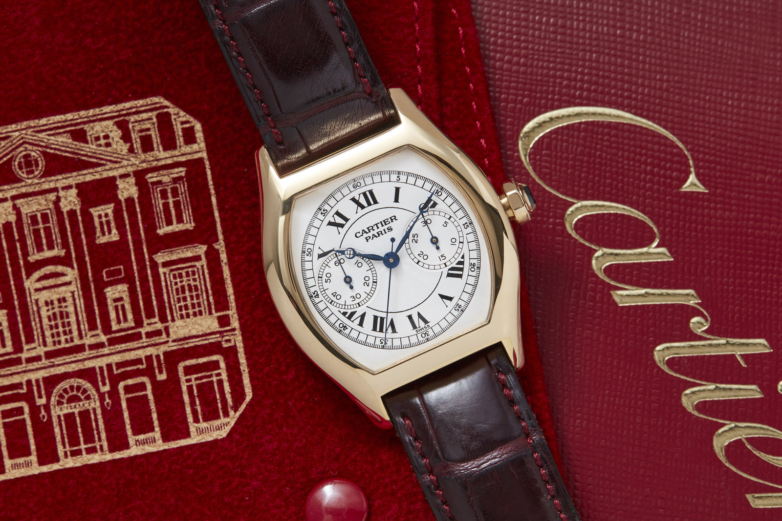A Cartier Tortue Monopusher Chronograph by Cartier with a brown leather strap rests on a red Cartier box, featuring a white dial, black Roman numerals, and two subdials.