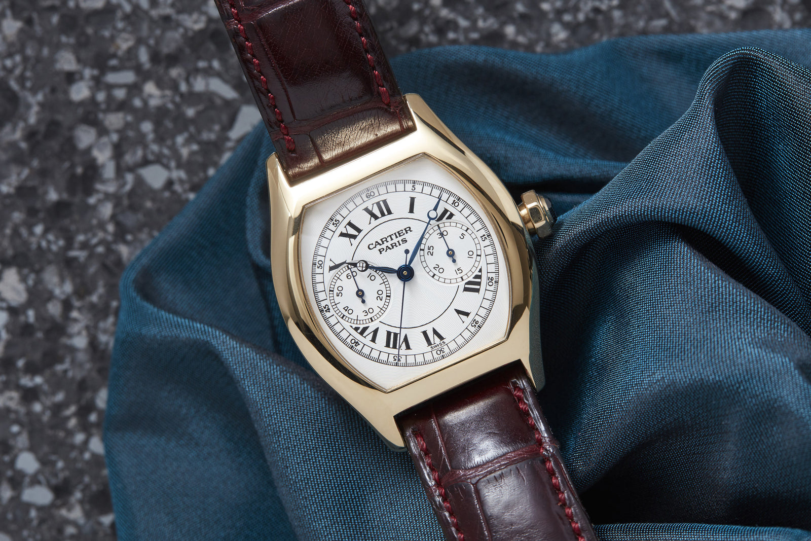 A Cartier Tortue Monopusher Chronograph, featuring a gold case, white Roman numeral dial, brown leather strap, and chronograph function, rests on a folded blue cloth over a speckled gray surface.