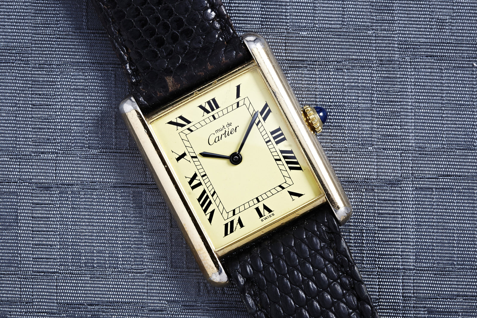 The Cartier Tank Manual Wind - Men's watch features a rectangular gold case, cream dial with black Roman numerals, and a black textured leather strap, showcased on a grey surface for an elegant, classic look.