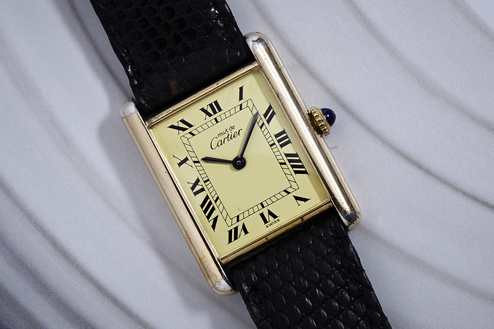 The Cartier Tank Manual Wind Men's watch features a cream dial with black Roman numerals and a black leather strap, combining classic elegance with refined military-inspired style. Displayed at 10:10 on a light textured surface.