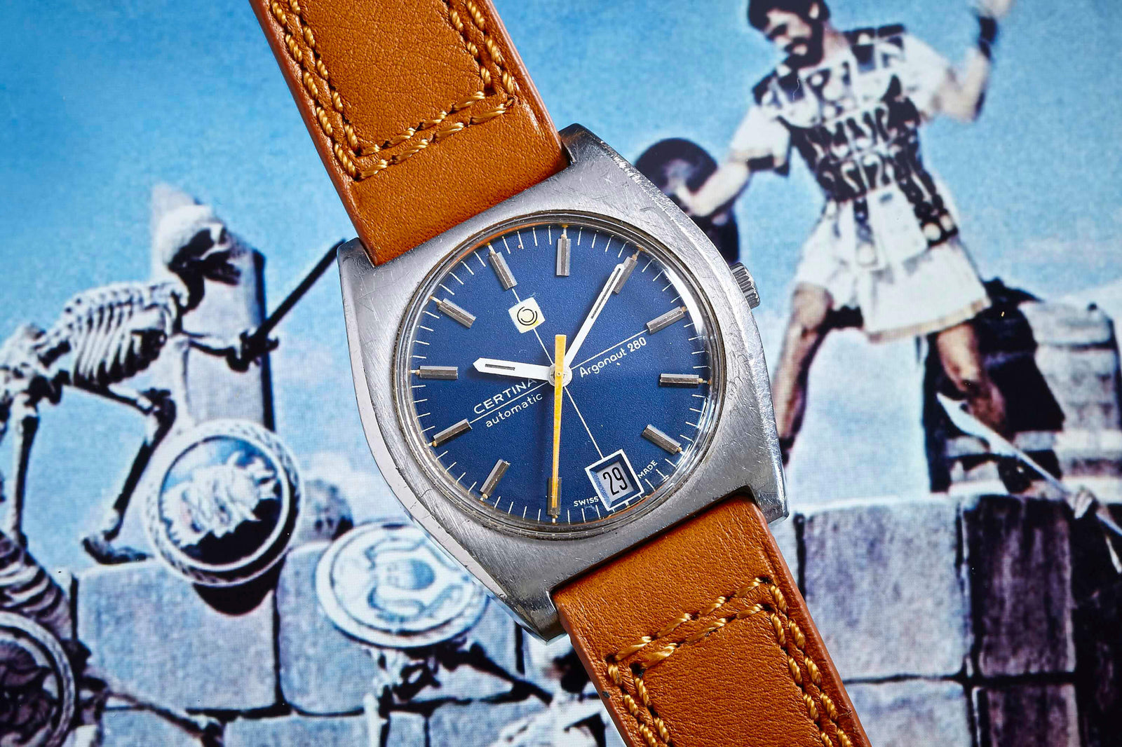 A Certina Argonaut 280 automatic wristwatch with a blue dial and brown leather strap is displayed against an image of ancient warriors and skeletons battling on stone steps.