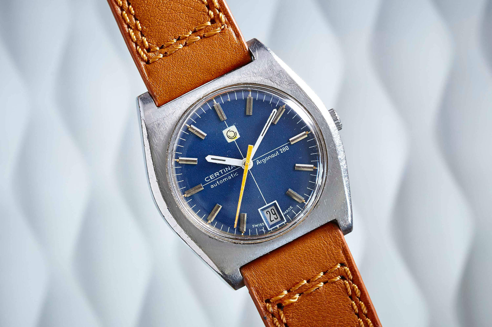 The Certina Argonaut 280 is a vintage automatic watch featuring a blue dial, date window, silver markers, yellow seconds hand, brown leather strap, and square silver case, displayed against a wavy white background.