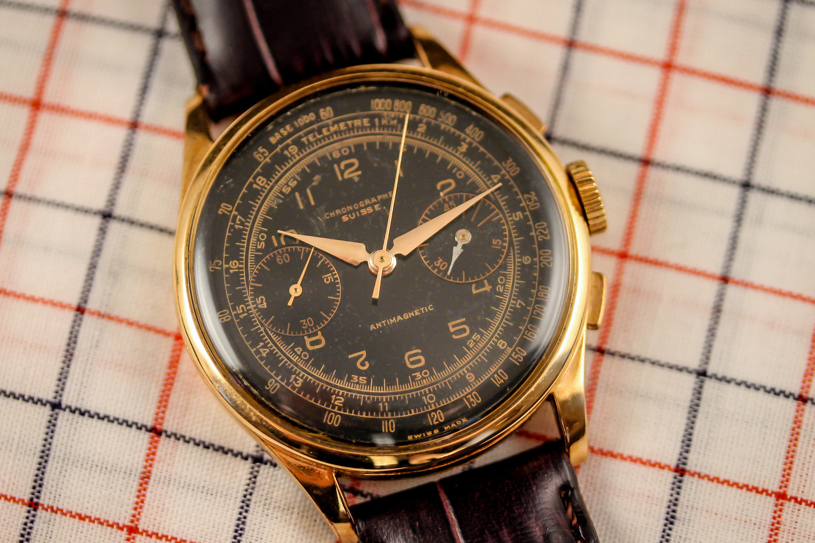 A Chronographe Suisse - Rare Black Dial vintage chronograph wristwatch with a brown leather strap rests on checkered fabric, celebrating the Swiss brand's heritage.