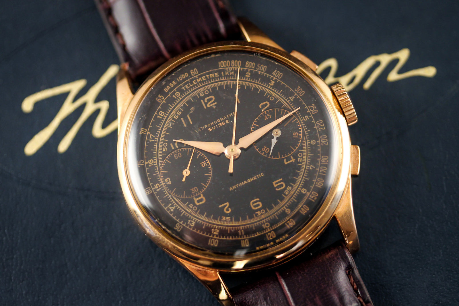 A Chronographe Suisse Rare Back Dial wristwatch with a brown leather strap and three subdials rests on a dark surface, with elegant gold script lettering in the background.