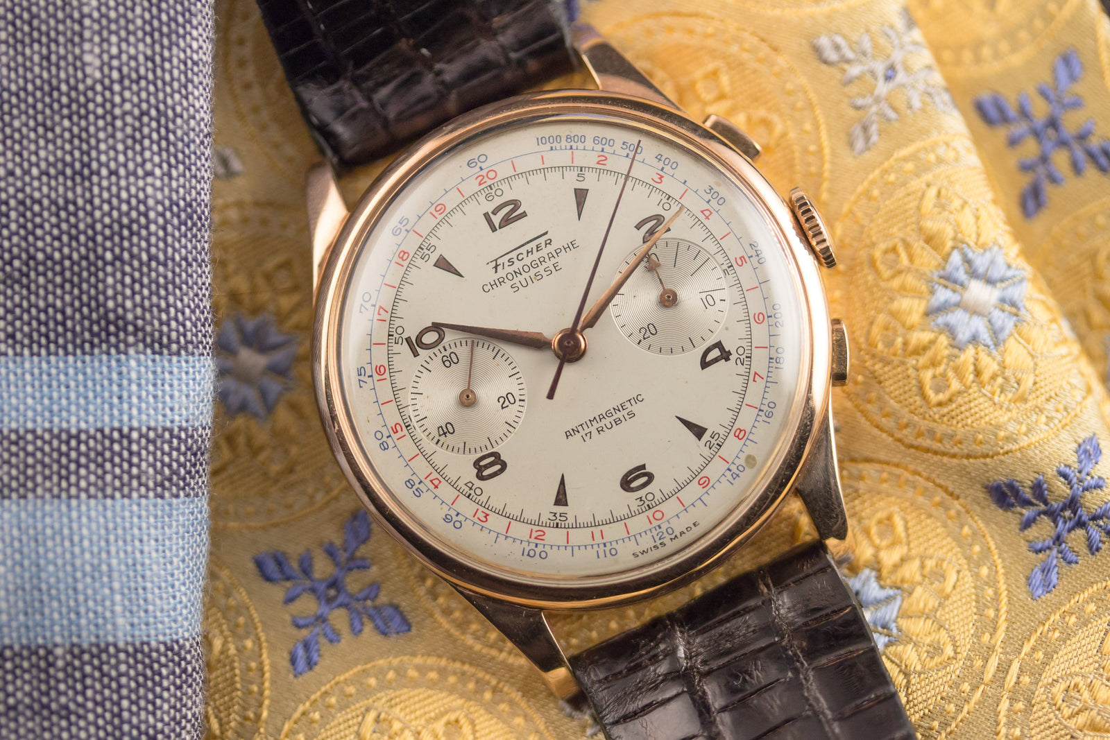 The Chronographe Suisse Pink Gold - Fischer Dial wristwatch by Chronographe Suisse rests on ornate gold and blue fabric, featuring a white dial with black Arabic numerals, subdials, and red and blue markings.
