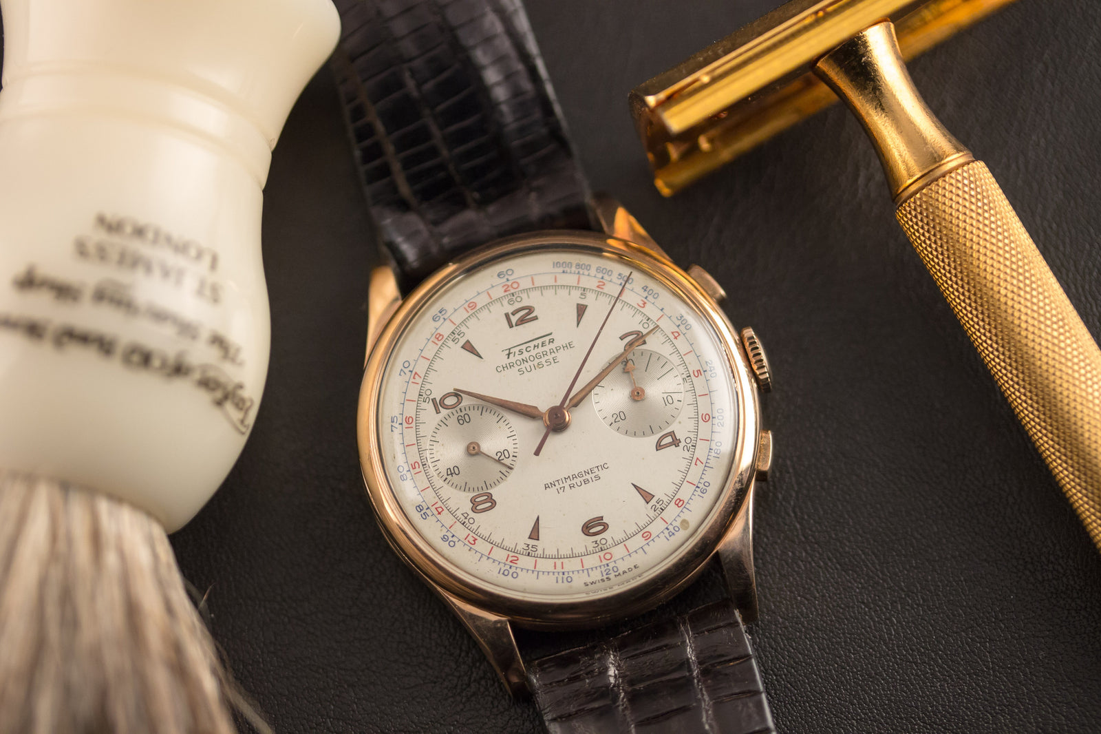 The Chronographe Suisse Pink Gold - Fischer Dial wristwatch rests beside a gold safety razor and white-handled shaving brush—a refined tribute to classic Swiss watchmaking by Chronographe Suisse.