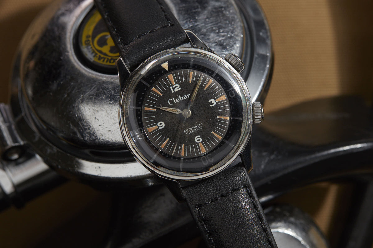 A Clebar Super Compressor wristwatch with a black leather strap and black dial sits on a metallic surface, evoking diving suit precision, with a circular Qualitron label in the background.