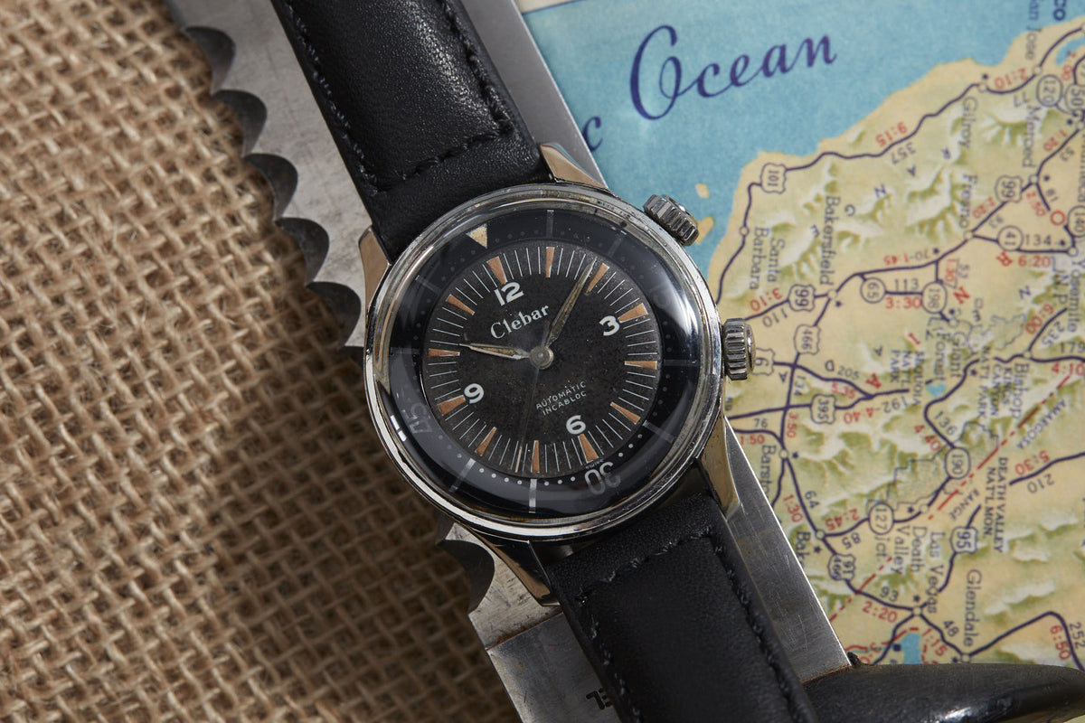 The Clebar Super Compressor by Clebar, with a black leather band, sits on a knurled metal blade atop burlap beside a vibrant map marked "Ocean," capturing the adventurous spirit of classic divers.