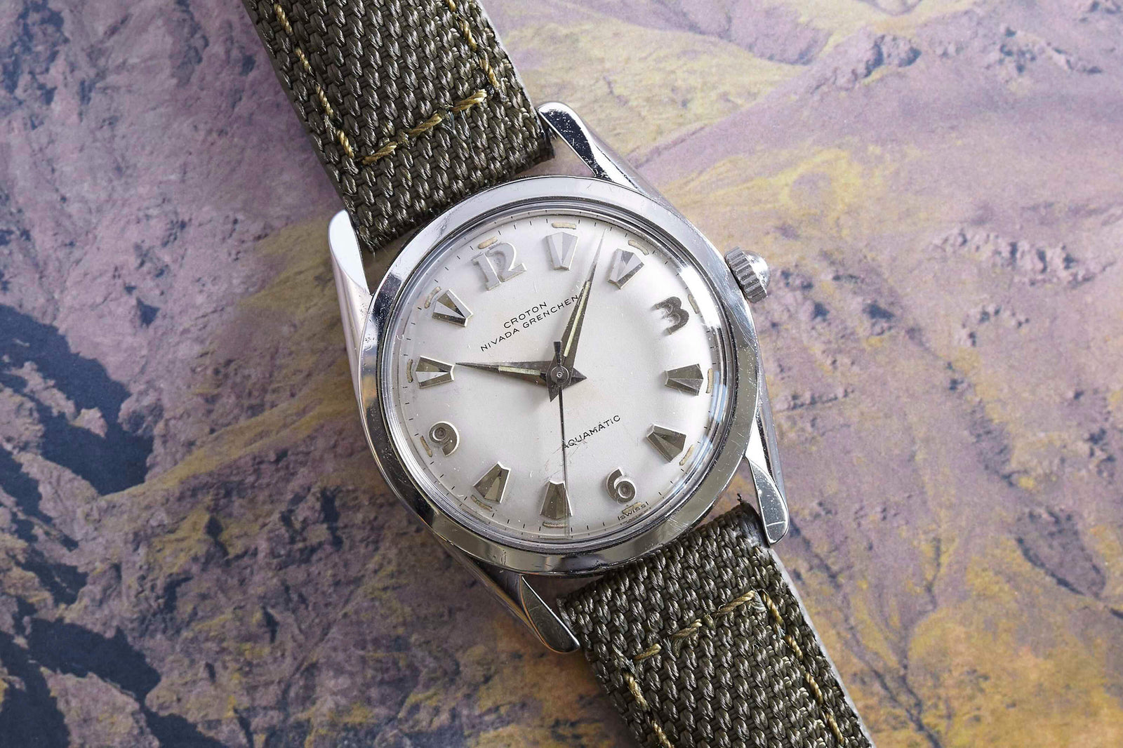 A vintage Croton Aquamatic by Croton featuring a silver case, olive green fabric strap, and silver numerals and hands, displayed on a map-like background with warm brown and yellow tones.