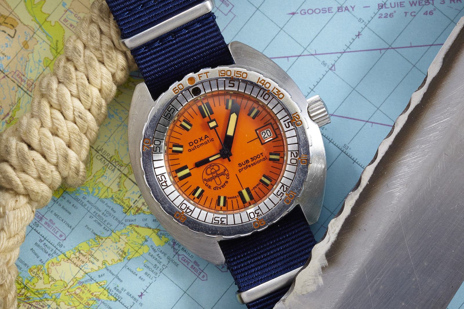 A DOXA Sub 300T Professional - US Divers Dial watch with an orange face and blue strap sits on a nautical map, displaying its signature uni-directional rotating bezel, next to coiled rope and a metal object.