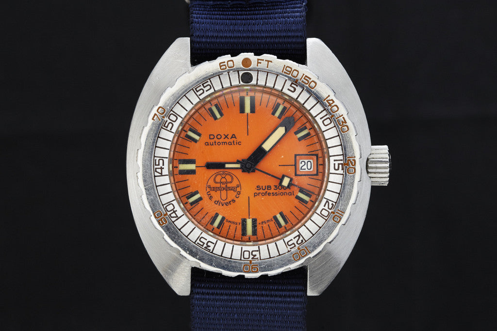 The DOXA Sub 300T Professional - US Divers Dial is a vintage diver’s watch with an orange dial, bold black and luminous markers, date at 3 o’clock, blue fabric strap, and a uni-directional rotating bezel for diving.