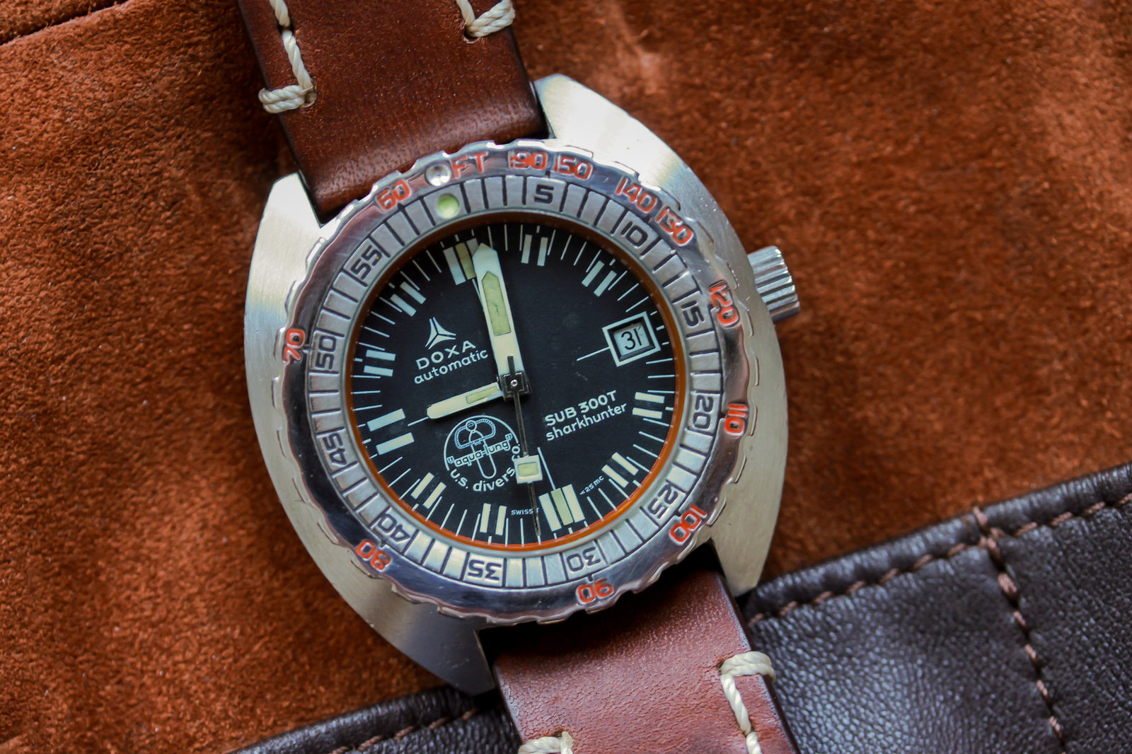 A close-up of the DOXA Sub 300T Sharkhunter Aqua Lung, a divers watch with a brown leather strap, black dial, date window, and orange accents, resting on a brown suede and leather surface.