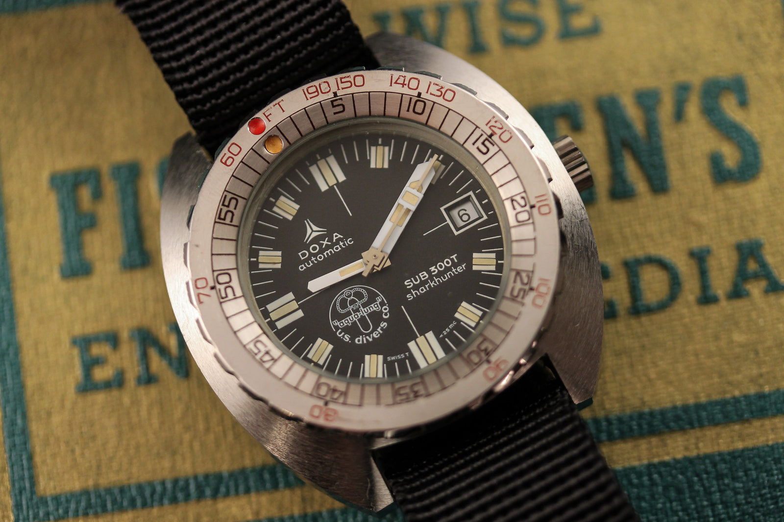 A close-up of the DOXA Sub 300T Sharkhunter Aqua Lung watch with a black strap, silver bezel, and black dial displaying 8:01 and the 6th, resting on a textured book cover.
