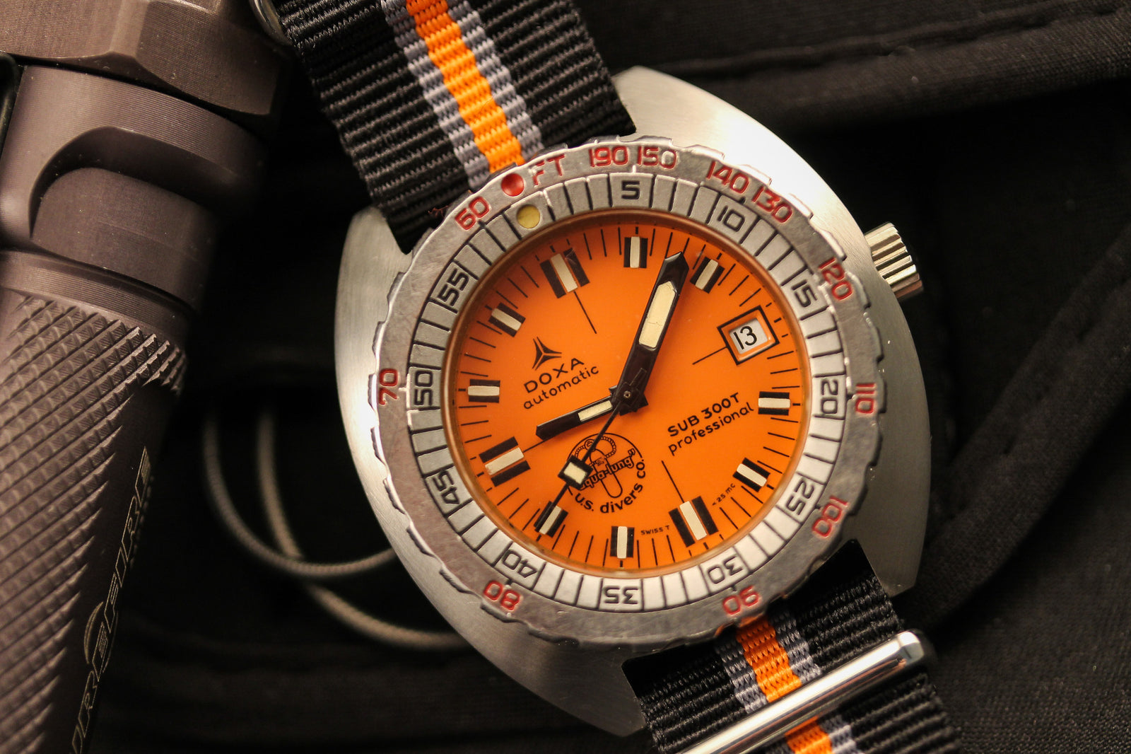 A close-up of the DOXA Sub 300T Professional Aqua Lung diver’s watch with an orange dial and bold date display, on a black and orange nylon strap beside a textured metallic object; features include bold markings and a Helium Release Valve.