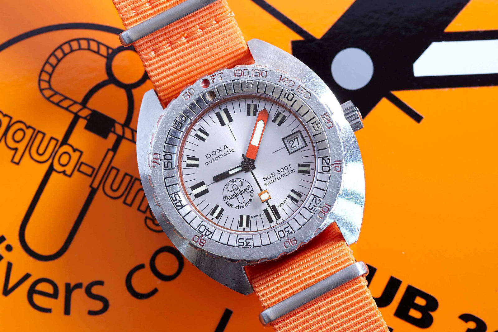 A DOXA Sub 300T Searambler dive watch with an orange strap lies on a bright orange Aqua-Lung box, showcasing a date window, bold black and orange hands, and the classic helium release valve.