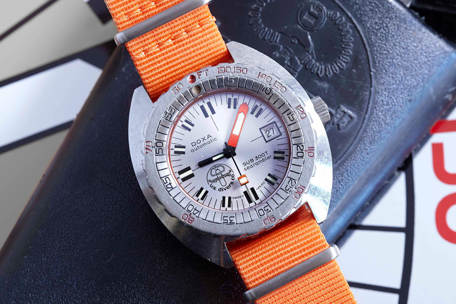 The DOXA Sub 300T Searambler, a silver dive watch by DOXA, features a white dial, bold hour markers, orange second hand and NATO strap, helium release valve, and shows the date as 21st. It rests on a black textured surface.